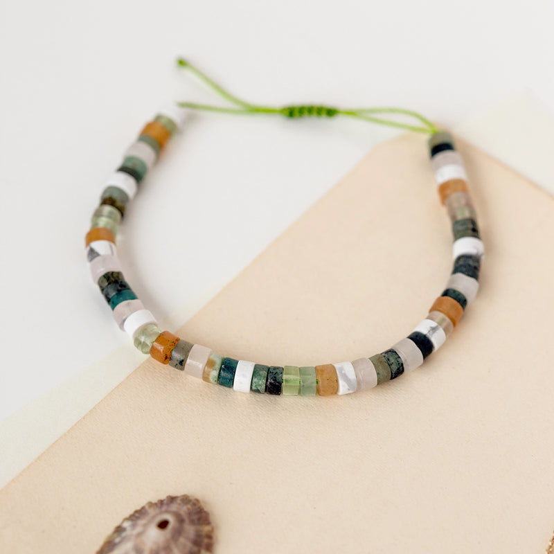 The seagrass cord bracelet from bluboho, featuring an olive-colored thread fastener and gemstone beads in green, white, black, and brown hues, is showcased on a cream-colored background. A small seashell is placed in the corner of the image.