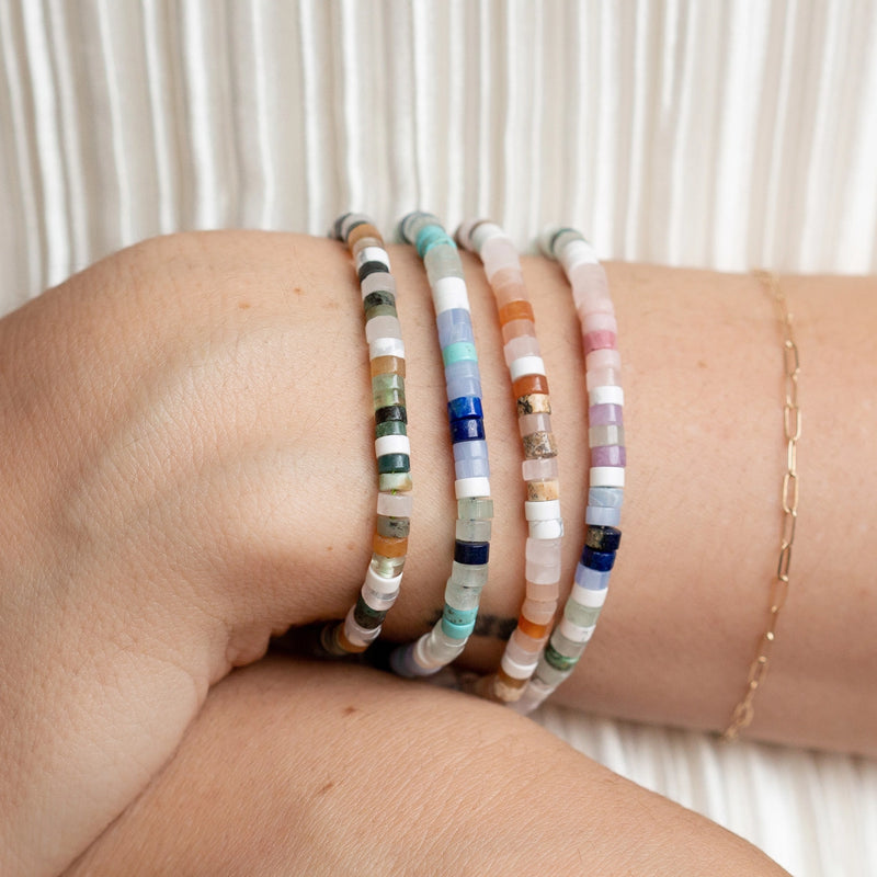 A close-up of a person's wrist showcases the Seagrass Cord Bracelet from Bluboho, adorned with an olive cord and colorful gemstone beads in shades of blue, pink, white, green, and orange. The backdrop is a white pleated fabric, and a delicate gold chain bracelet can be seen partially on the same arm.