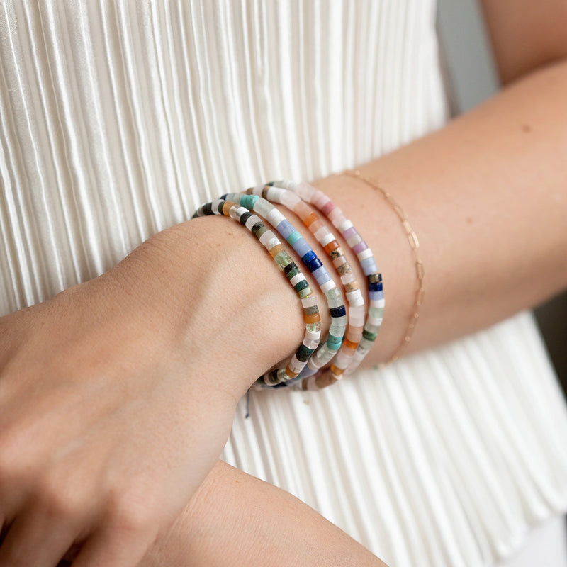 A person wears multiple beaded bracelets with various colored beads on their wrist, including the "rainbow bead bracelet" from bluboho, which features gemstone beads on a beige cord. Alongside these, they also sport a thin, delicate gold chain bracelet. They are dressed in a white pleated top. The image focuses on the bracelets and the lower part of their arm.