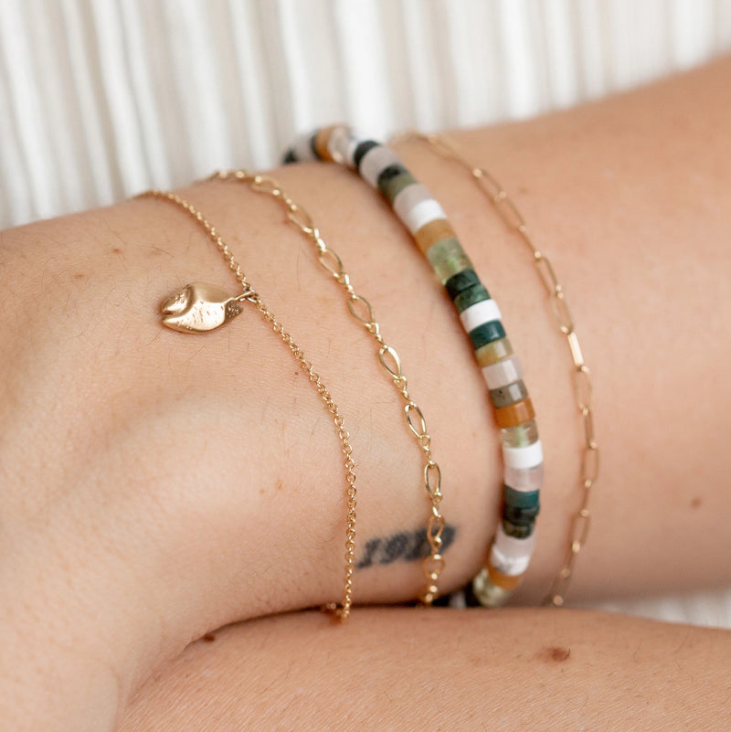 Close-up of a wrist adorned with five different bracelets. The collection includes delicate gold chains, one featuring a small heart charm, and the bluboho seagrass cord bracelet, which combines olive cord with muted earth-toned gemstone beads. A small tattoo is barely visible beneath the gold chain bracelets.