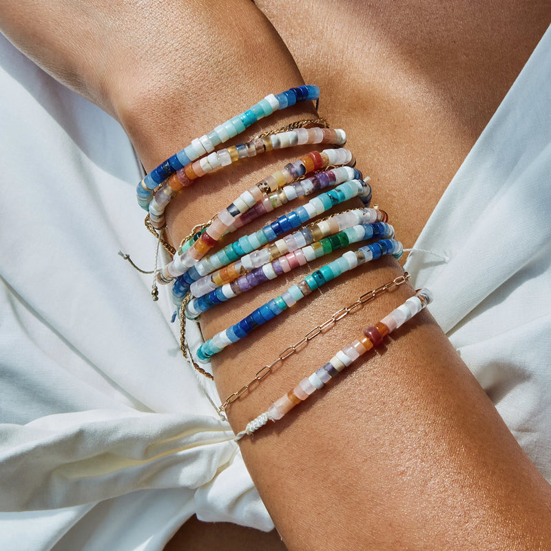 Close-up of a person’s wrist adorned with multiple colorful beaded bracelets, including the *ocean blu bead bracelet - blue cord with gemstone beads* by *bluboho*. The bracelets feature a variety of beads in colors like blue, pink, white, orange, and gold. The person is wearing a light-colored garment, partially visible in the background.