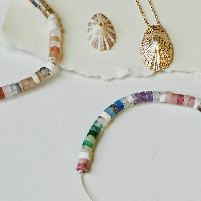 A photograph features a gold seashell pendant necklace, a small seashell, and two bluboho rainbow bead bracelets with beige cords and gemstone beads in pastel colors. The beads form curved lines in the image, and the items are placed on a white surface with a torn piece of white paper in the background.