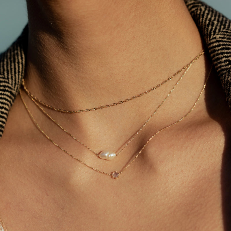 Close-up of a person's neck displaying three delicate gold necklaces. The shortest necklace is a thin chain, the middle one is the bluboho sofia perla necklace - a 14k yellow gold necklace featuring a small white pearl, and the longest one holds a small, light-colored gemstone. The person is wearing a textured fabric jacket.