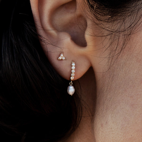 Close-up of a person's ear wearing two earrings. The first is a small gold stud with three tiny pearls, placed on the upper part of the earlobe. The second is the "inner light diamond pearl earring" by bluboho, crafted from 14k yellow gold and adorned with small diamonds and a single dangling white pearl, located on the lower lobe.