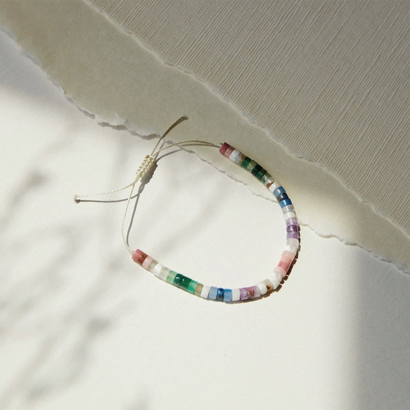 The rainbow bead bracelet from bluboho, featuring gemstone beads on a beige adjustable cord, is displayed on a textured off-white background. This exquisite piece showcases a mix of pastel and vibrant shades. Soft sunlight casts gentle shadows across the scene.