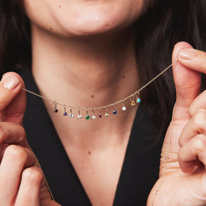 A person dressed in black is holding a delicate bluboho 10k yellow gold necklace adorned with the Serendipity July Birthstone Charm featuring a captivating ruby. The necklace is held in both hands, stretched out to display various gemstone charms in shades of blue, red (including the ruby), green, purple, and clear.