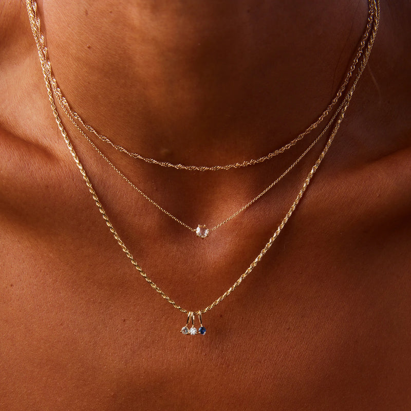 Close-up image of a person with a bronzed complexion wearing three layered gold necklaces. The top necklace is thin with a small, circular pendant. The middle necklace also has a small, round pendant. The bottom necklace features the serendipity july birthstone charm in 10k yellow gold, adorned with a ruby, from bluboho.