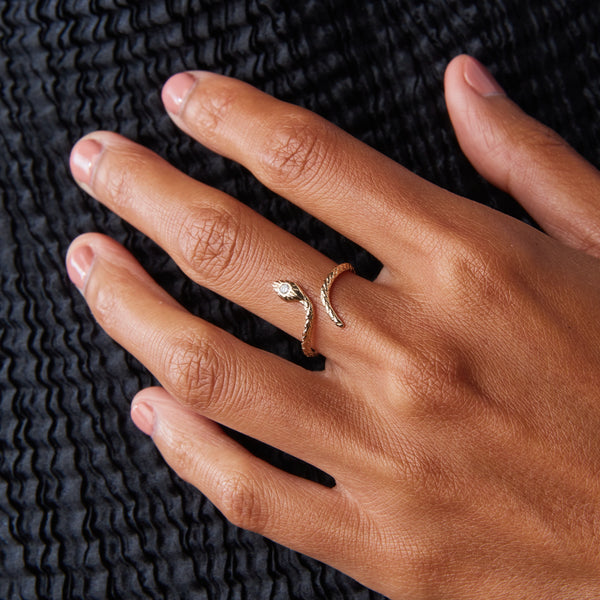 Close-up of a hand adorned with the Serpent Revival Diamond Cuff Ring by Bluboho, featuring 14k yellow gold with a small diamond on its head. The hand rests gracefully on a textured, dark background, while the ring elegantly wraps around the finger, beautifully complementing the warm tone of the skin.