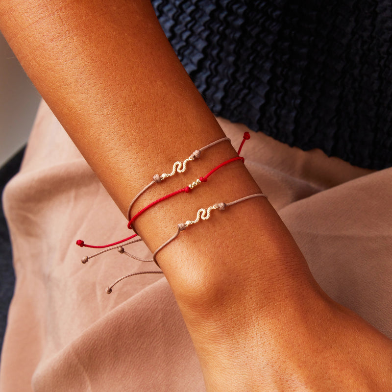 Close-up of a person's wrist adorned with three delicate bracelets. Two of the bracelets have beige cords, while the third features the abacus contemplation cord bracelet in red with 10k yellow gold from bluboho. Each bracelet is accented with a wavy, gold-colored charm. The person is dressed in textured, dark-colored fabric.