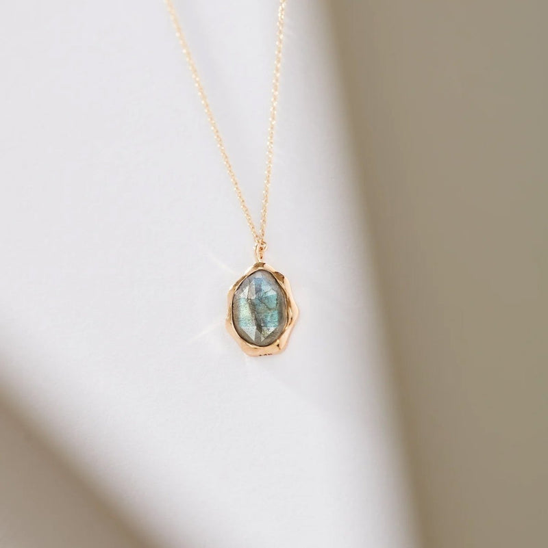 The labradorite mood necklace by bluboho is showcased against a white backdrop, highlighting its delicate gold chain. The pendant boasts an oval-shaped, iridescent labradorite gemstone set within a gold bezel, radiating a soft glow with subtle shades of blue and green.