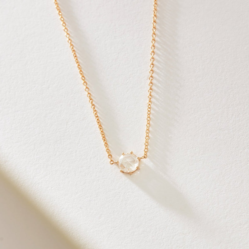 A delicate extra large supernova necklace from bluboho, crafted in 14k yellow gold, is displayed on a white surface. The necklace features a small, round, faceted crystal or gemstone pendant set in a simple prong setting, centered on the chain. The shadows suggest the necklace is resting in soft, natural light.