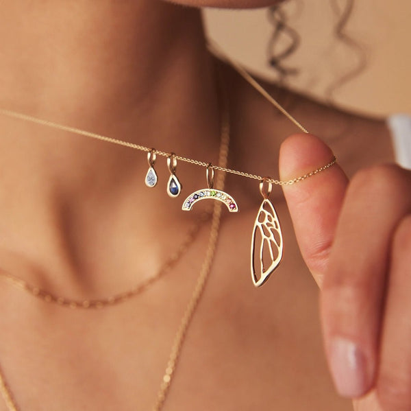 A person adorned with a gold necklace from bluboho is pictured, showcasing four distinct charms. These include a teardrop-shaped gem from the pear diamond April mood birthstone charm in 10k yellow gold, a multicolored arc, a bee wing, and another teardrop-shaped gem. The person's hand delicately holds the chain, drawing attention to the exquisite charms.