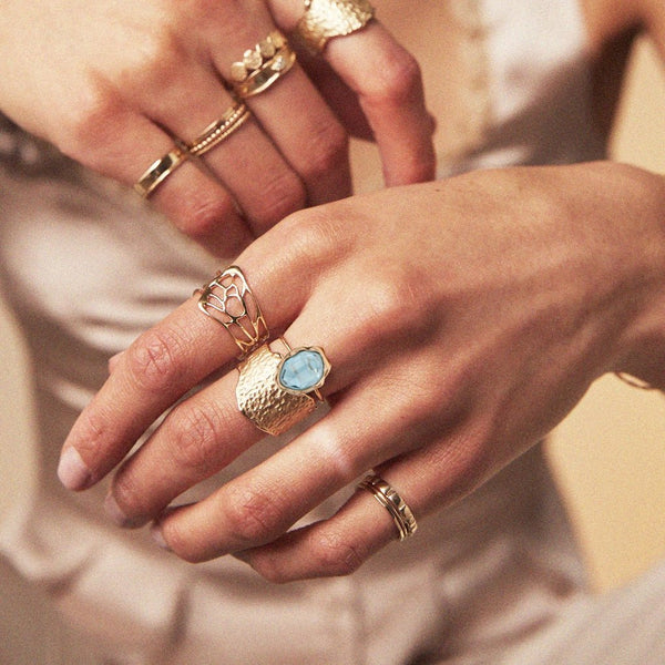 Close-up of a person's hands adorned with rings. One hand features a bluboho "london blue topaz mood ring" crafted from 14k yellow gold, showcasing a striking blue topaz stone. The other fingers display an assortment of gold rings with varying designs, some intricately detailed and others as simple bands. The softly blurred background accentuates the beauty of the jewelry.