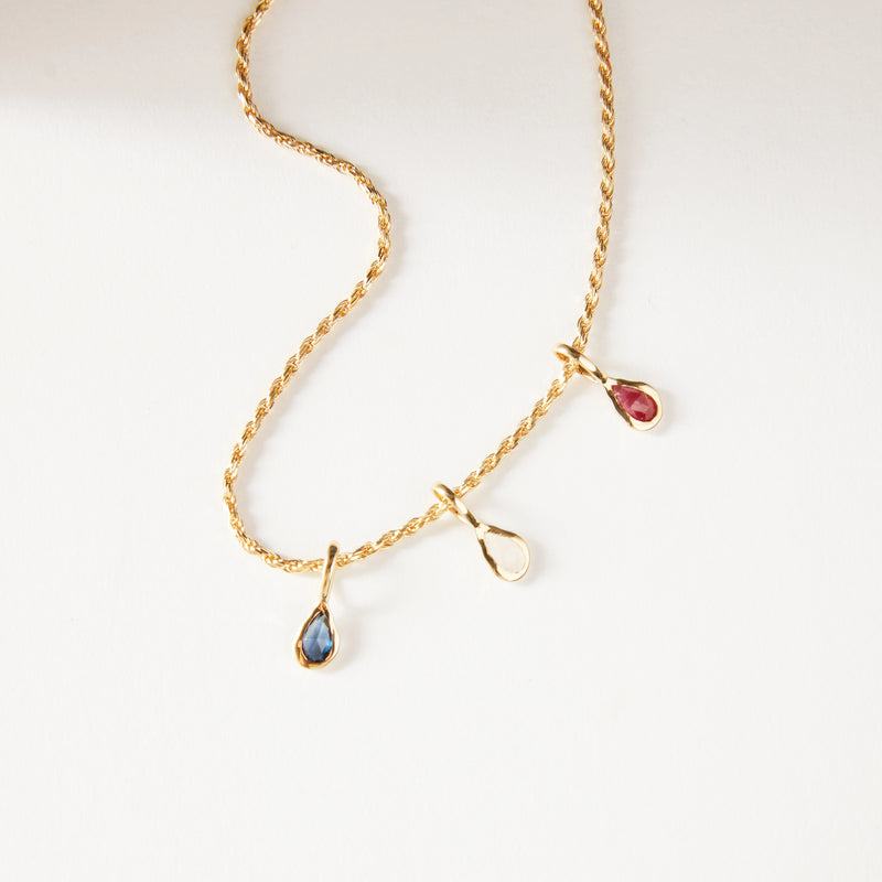 A 10k yellow gold twisted chain necklace by bluboho with three teardrop-shaped gemstone pendants, featuring blue turquoise, clear, and red gemstones, each encased in a gold setting. Elegantly displayed on a white background.