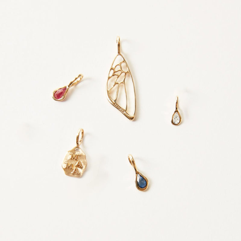 A captivating array of five 10k yellow gold pendants from bluboho showcased on a white background. Each pendant boasts unique designs: one featuring a butterfly wing pattern, another adorned with a red teardrop gem, the third with a clear teardrop gem, an irregularly textured piece, and the fifth highlighting the elegant pear diamond April mood birthstone charm.