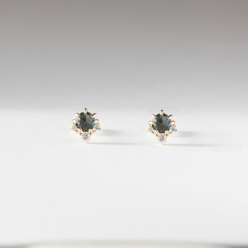 Introducing the Full Moon Glow Diamond London Blue Topaz Earrings by bluboho: These elegant stud earrings feature round, blue topaz gemstones set in a 14k yellow gold prong setting with small, clear diamond accent stones. They are showcased on a neutral, light gray background, positioned symmetrically and viewed from a front perspective.