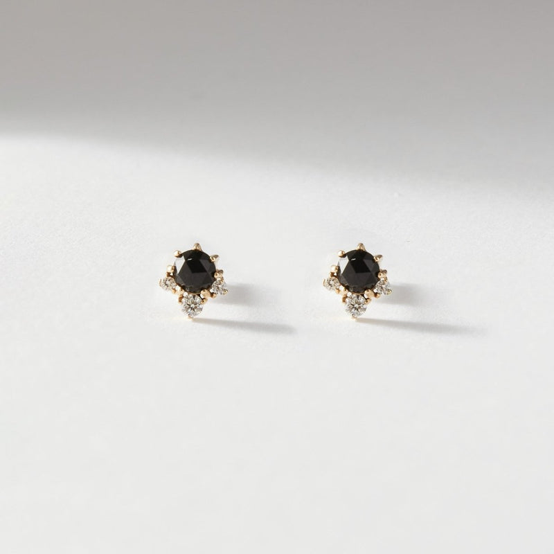 A pair of Bluboho Full Moon Glow Diamond Black Onyx Earrings - 14k yellow gold, black onyx, and diamonds is photographed on a light-colored, textured surface. These elegant earrings feature black gemstones set in gold with small white stones intricately arranged around the black onyx.