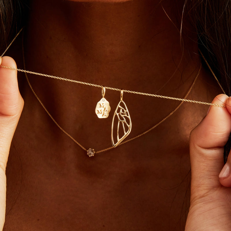 A woman holds a delicate gold necklace with two pendants close to her chest. One pendant is a teardrop shape with "MAMA" engraved on it, and the other is bluboho's intricate butterfly wing charm made of 10k yellow gold. Another simple gold chain with a small round pendant rests below it.