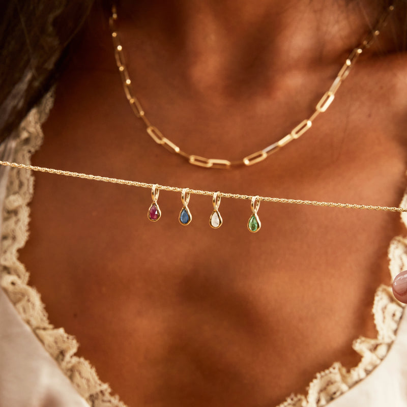 A person wearing a gold chain necklace with elongated rectangular links holds a delicate chain featuring four teardrop-shaped pendants in different colors: red, blue, clear, and green. The person is dressed in a light-colored, lace-trimmed garment. One of the pendants is the bluboho pear amethyst february mood birthstone charm - 10k yellow gold.