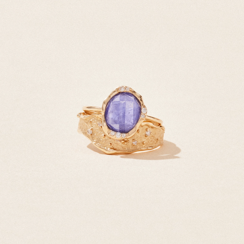 The 14k yellow gold *tanzanite mood ring* by bluboho features a textured band with small embedded diamonds and showcases a large, oval blue tanzanite gemstone as the centerpiece. The intricate details of the ring create a striking and elegant appearance.