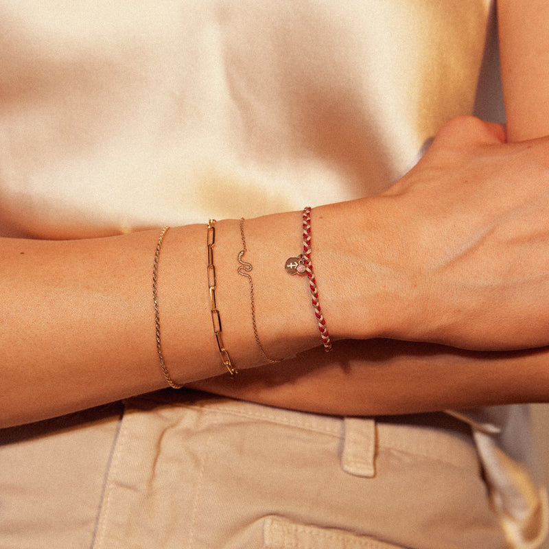 A person wearing a white outfit showcases their forearm adorned with four different bracelets. The bracelets include a thin gold chain, a thicker linked gold chain with a small snake charm, and a red braided bracelet with a small charm from bluboho's aries zodiac fire element cord bracelet - 10k, opal, cord. The background is softly lit.