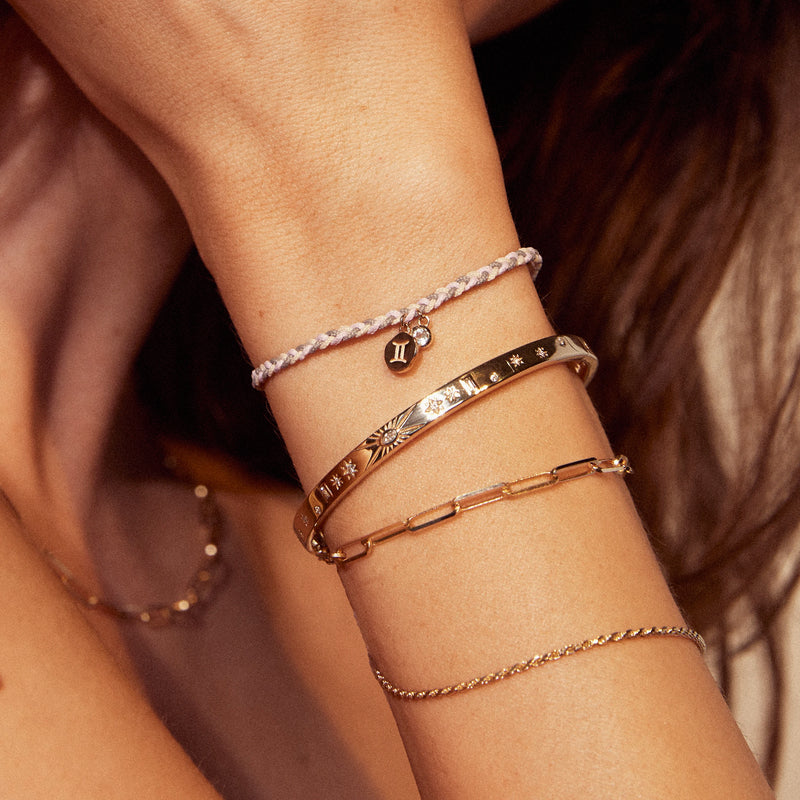 Close-up of a person's wrist adorned with various bracelets: a delicate chain bracelet, a bluboho libra zodiac air element cord bracelet - 10k yellow gold, aquamarine, cord, a gold bracelet with celestial engravings, and a gold link bracelet. The person also wears a necklace with a subtle chain.