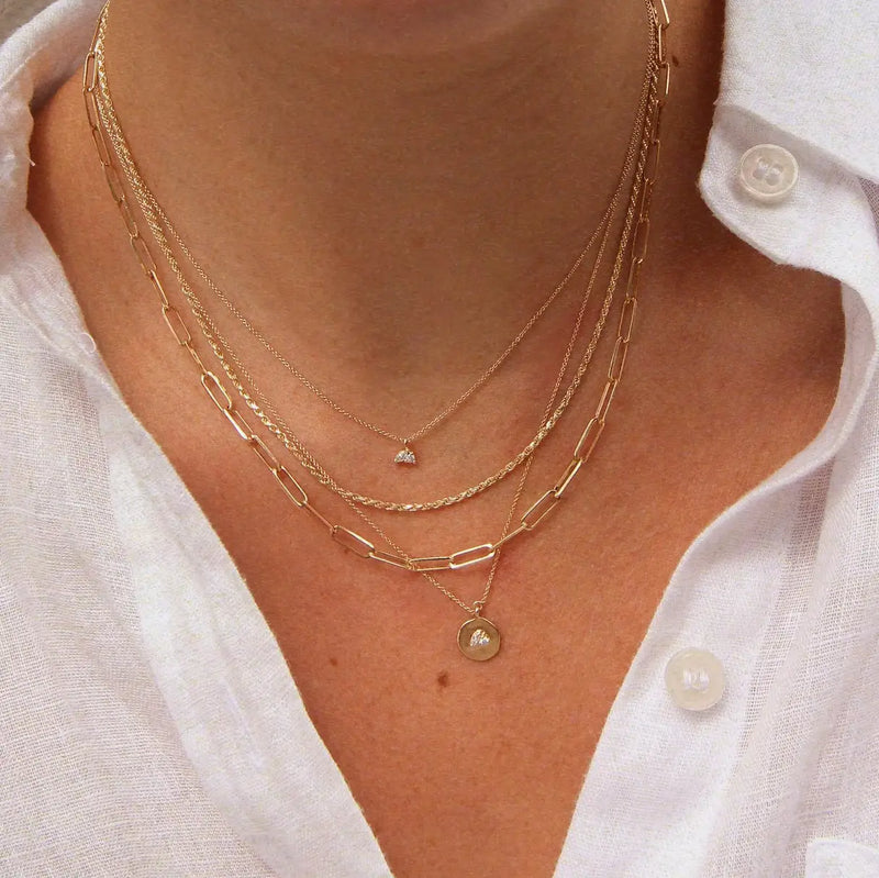 A close-up of a person wearing a white button-down shirt, showcasing three layered gold necklaces. One necklace is the "kindred spirits lean on me medallion necklace - 14k yellow gold, diamonds" from bluboho, another features a delicate chain design, and the third has a circular pendant with an engraved letter.