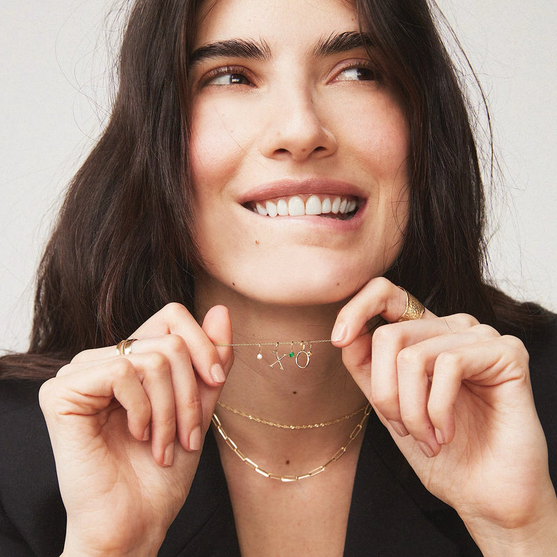 A woman with long dark hair smiles and looks up while holding a delicate bluboho letter charm - 10k yellow gold near her mouth. She wears multiple layers of gold necklaces and rings, and a black top. She's expressing a playful and joyful demeanor against a plain background.