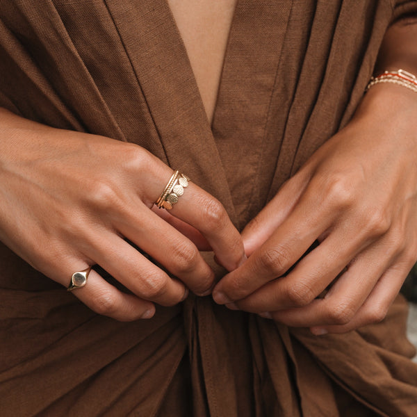 Close-up of a person wearing a brown, draped top, showing their hands adorned with various rings and bracelets. The focus is on the hands, showcasing gold and silver jewelry, including a bluboho abacus disk ring - 14k yellow gold and stacked rings with small charms.