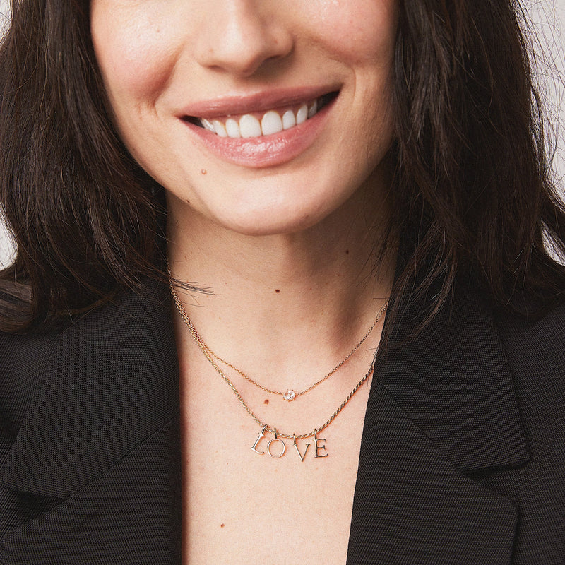 A woman with long, dark hair is smiling. She is wearing a black blazer and two necklaces. One necklace is a simple gold chain, while the other is a bluboho letter charm - 10k yellow gold with the word "LOVE" spelled out in letters. The background is plain and light-colored.