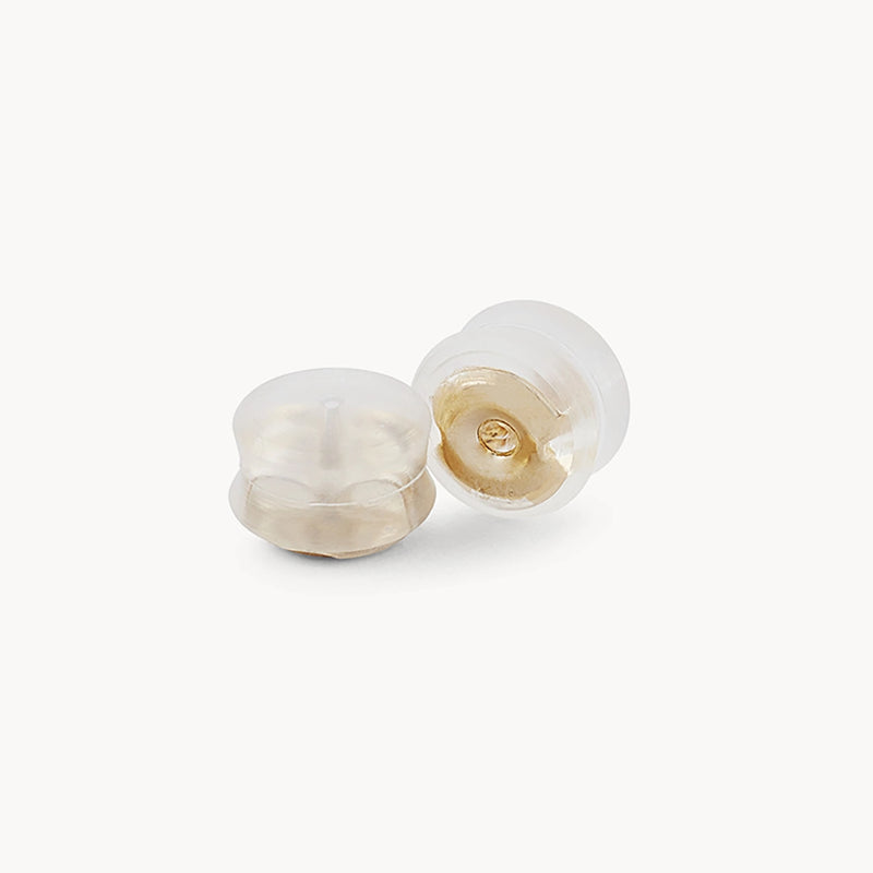 Two transparent silicone earring backs from bluboho's Larger Tree Bark Stability Hoop - 14k Yellow Gold collection are shown against a white background. One earring back stands upright, while the other leans to the side, displaying its hollow interior.