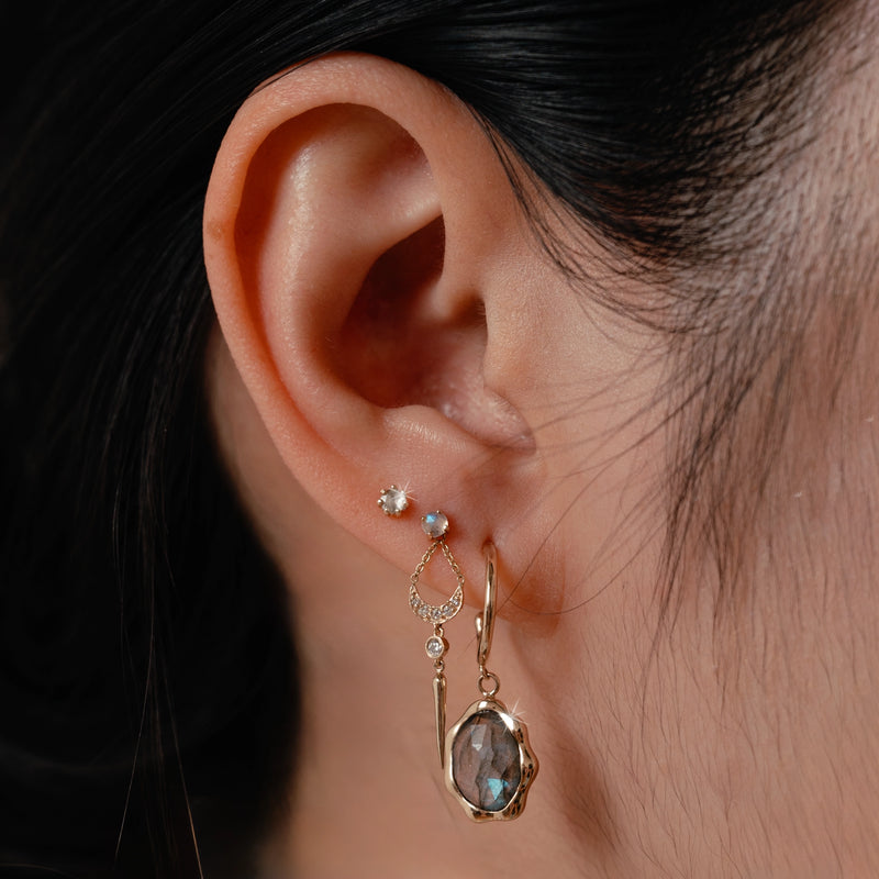 Close-up of a person's ear adorned with multiple earrings. The ear features a small hoop earring with a blue stone, a teardrop-shaped earring with a gem, and the bluboho nova earring - 14k yellow gold, white diamond. The person has dark hair pulled back.