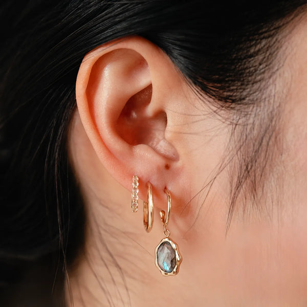 Close-up of a person's ear adorned with three gold hoop earrings from bluboho. The two smaller hoops are plain, while the larger labradorite mood hoop earring, crafted in 14k yellow gold, features a dangling, irregularly-shaped labradorite gemstone. The background is out of focus, emphasizing the jewelry.