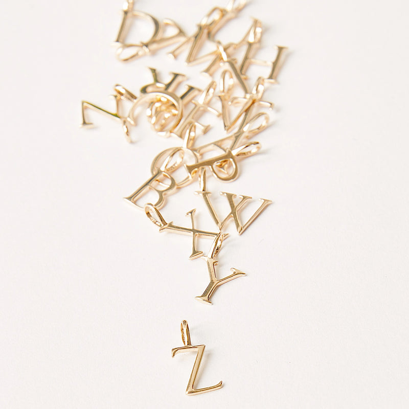 A collection of bluboho letter charm - 10k yellow gold scattered on a white surface. The letters appear randomly placed, starting from S at the top left and ending with a Z at the bottom right.