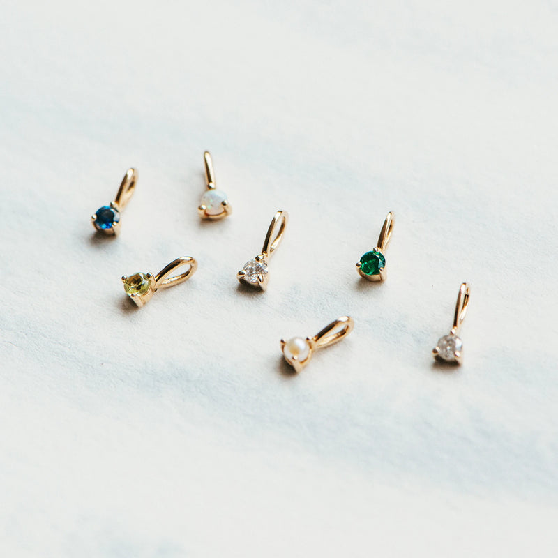 A set of seven small 10k yellow gold earring charms by bluboho, featuring a variety of gemstones such as blue, green, white, and sparkly clear stones, neatly arranged on a light marbled surface. The collection includes the stunning serendipity July birthstone charm with ruby.