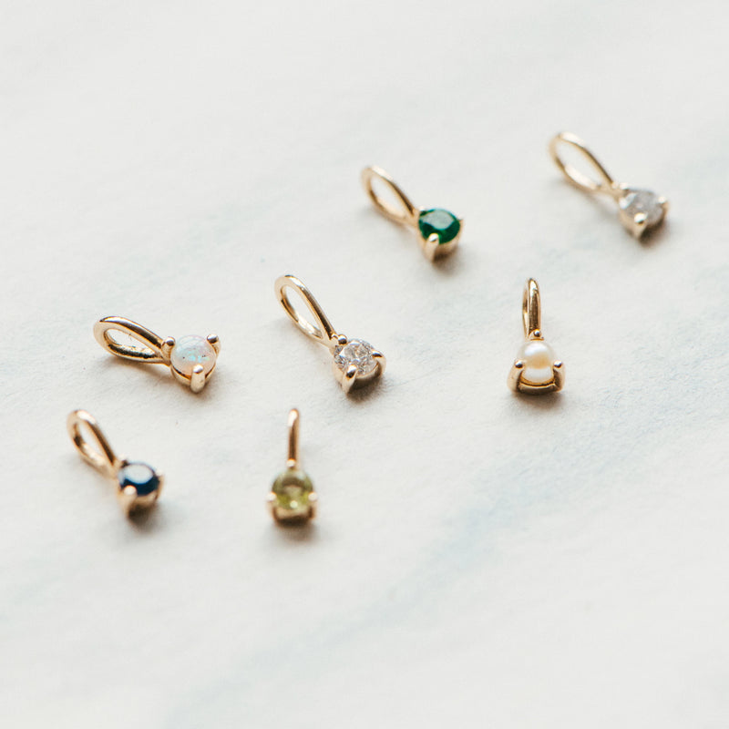 A close-up of seven small **serendipity August birthstone charms** by **bluboho**, crafted in 10k yellow gold, each uniquely adorned with a different gemstone. The stones exhibit a variety of colors, including blue, green (featuring peridot), white, and pearl. They are displayed in a loose grouping on a light-textured surface.