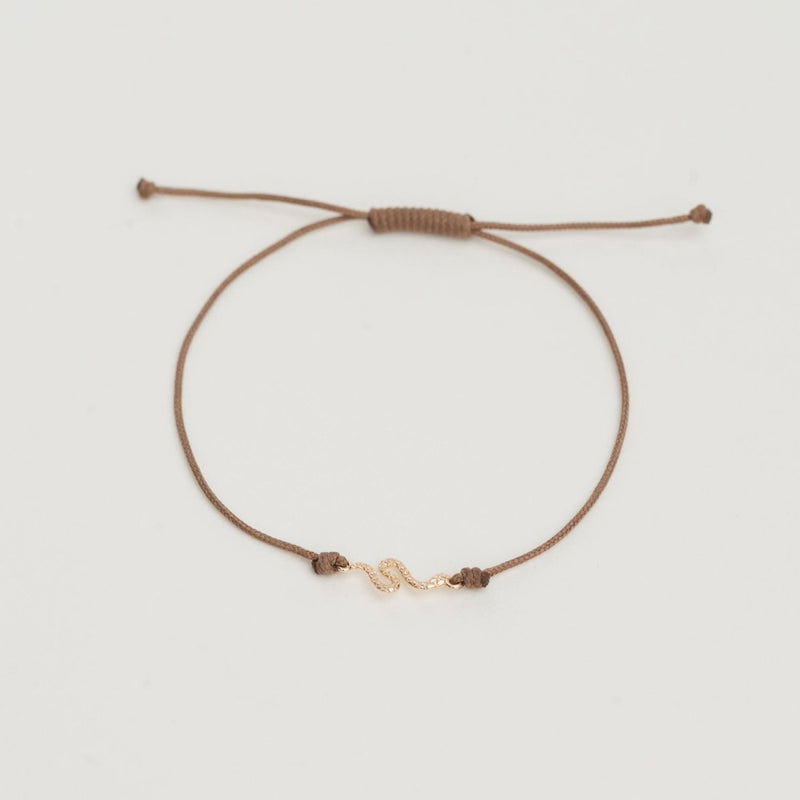 Introducing the bluboho Serpent Contemplation Cord Bracelet: a minimalist taupe string bracelet featuring an adjustable knot closure. The centerpiece is a small, 10k yellow gold squiggly design set against a plain white background.