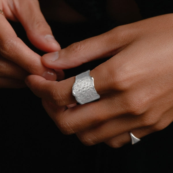 A close-up of a person's hands with slightly tan skin, showcasing the "sweetness of honey ring silver" in sterling silver by bluboho on one finger. The person's other hand is adjusting the ring while the dark background enhances the visibility of both the hands and the eye-catching statement ring.