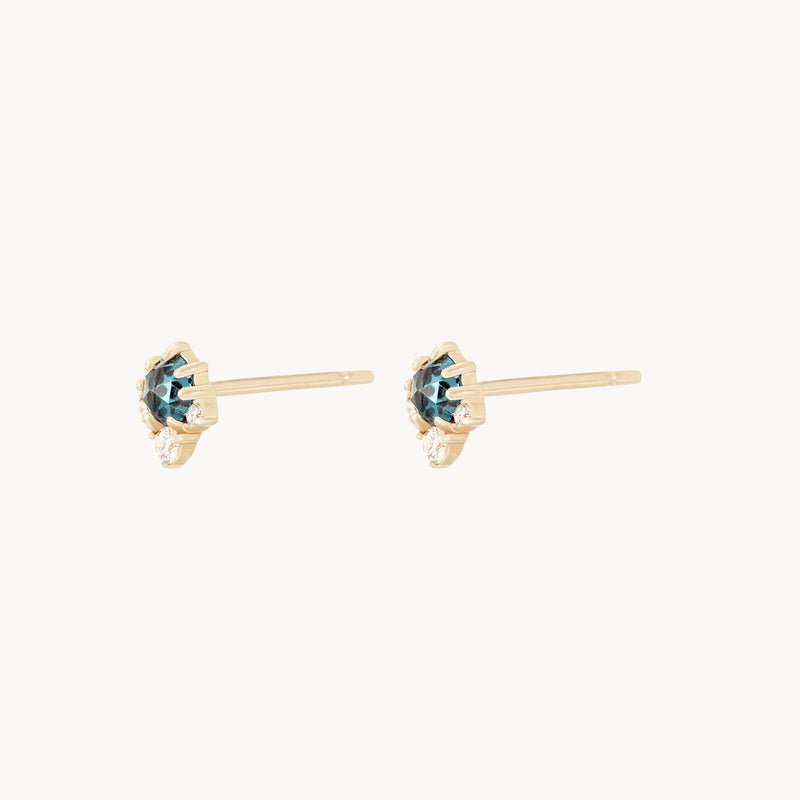 The "full moon glow diamond London blue topaz earrings" from bluboho are a pair of delicate 14k yellow gold stud earrings featuring mesmerizing blue topaz gemstones at the center, surrounded by sparkling diamonds. The earrings boast a simple post-back design and are displayed against a plain white background.