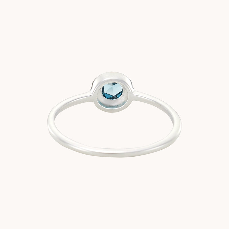 The full moon london blue topaz ring in sterling silver by bluboho showcases a minimalist design with a small, round blue gemstone set in a bezel setting. The thin band gently curves around the stone, accentuating its simple yet elegant appearance. The ring is displayed against an off-white background, highlighting its delicate beauty.