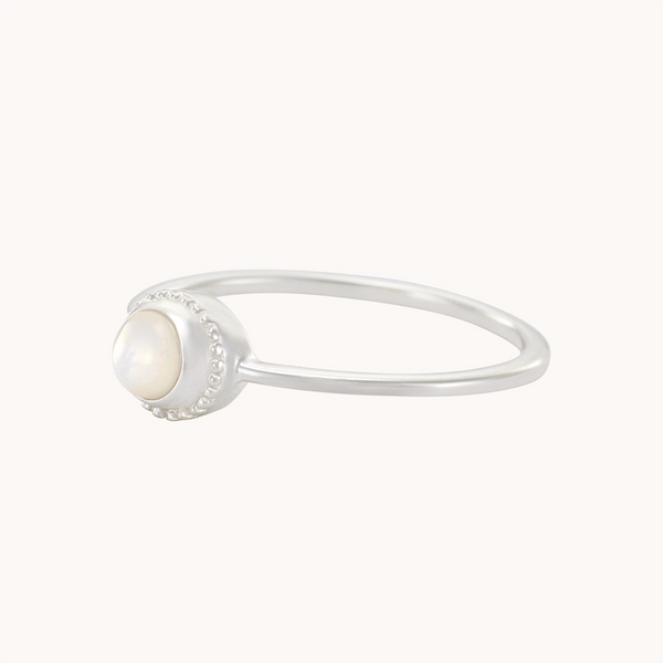 The Full Moon Light Mother of Pearl Ring Silver by Bluboho is a delicate silver ring featuring a small round mother of pearl stone set at the center. The stone exhibits a subtle iridescence, and the vintage-inspired setting boasts a beaded edge encircling the stone. The thin, polished band enhances the ring's elegant appearance.