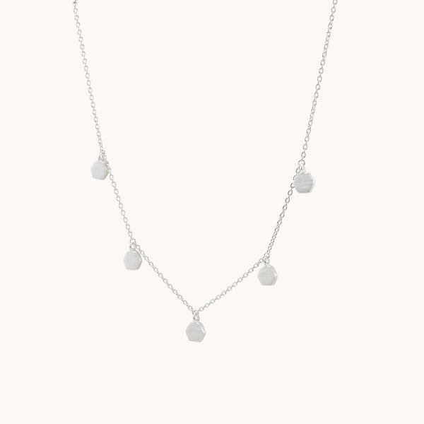 The honeysuckle necklace silver by bluboho is a delicate sterling silver piece that features a simple chain decorated with five evenly spaced small hexagonal pendants. Its minimalistic design evokes a modern and elegant style, beautifully highlighted against a plain, light-colored background to showcase its intricate details.