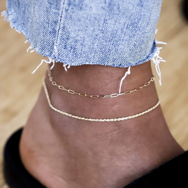 Close-up of an ankle adorned with two delicate "Infinite inseparable anklets" in 14k yellow gold by bluboho. The ankle is partially covered by the frayed hem of light blue, ripped jeans. The background is light brown, likely a wooden floor, and the person is wearing black shoes.