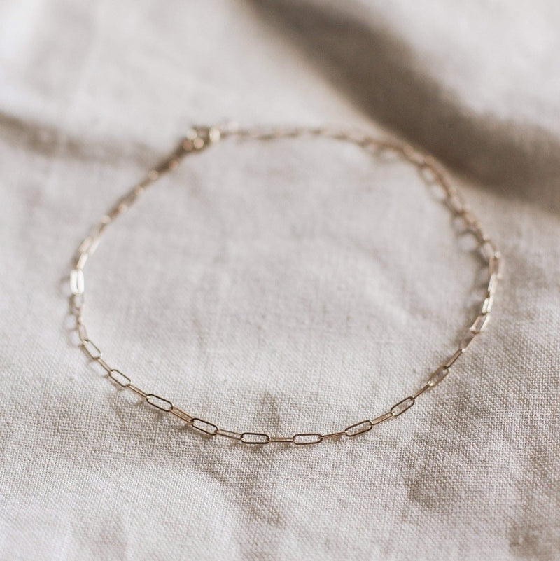 A delicate Infinite Inseparable Anklet by bluboho, crafted from 14k yellow gold with interlocking oval links, is laid out on a softly textured, light-colored fabric. The simplicity and elegance of the jewelry contrast beautifully with the neutral background.