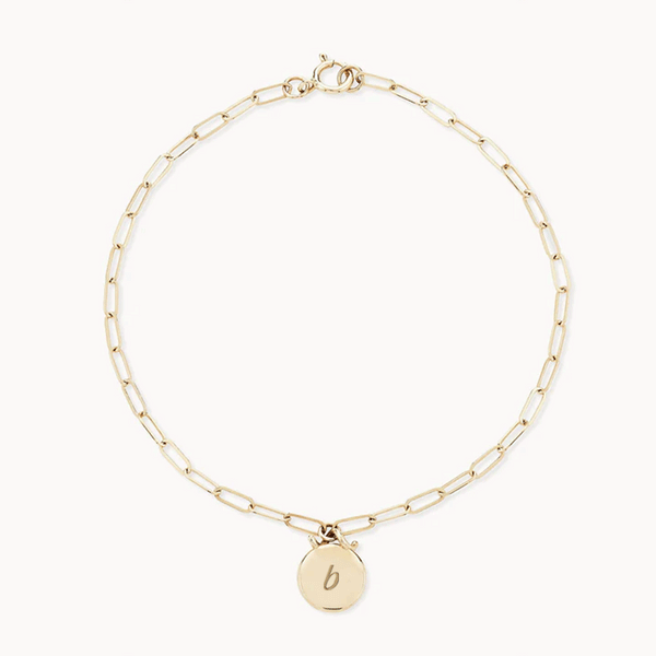 A delicate gold chain bracelet featuring elongated, rectangular links. It has a circular charm with the letter "b" engraved on it, attached via a small loop. The bluboho Imprint Forest Bracelet - 14k Gold, Engravable is laid out in a circular shape on a plain white background.