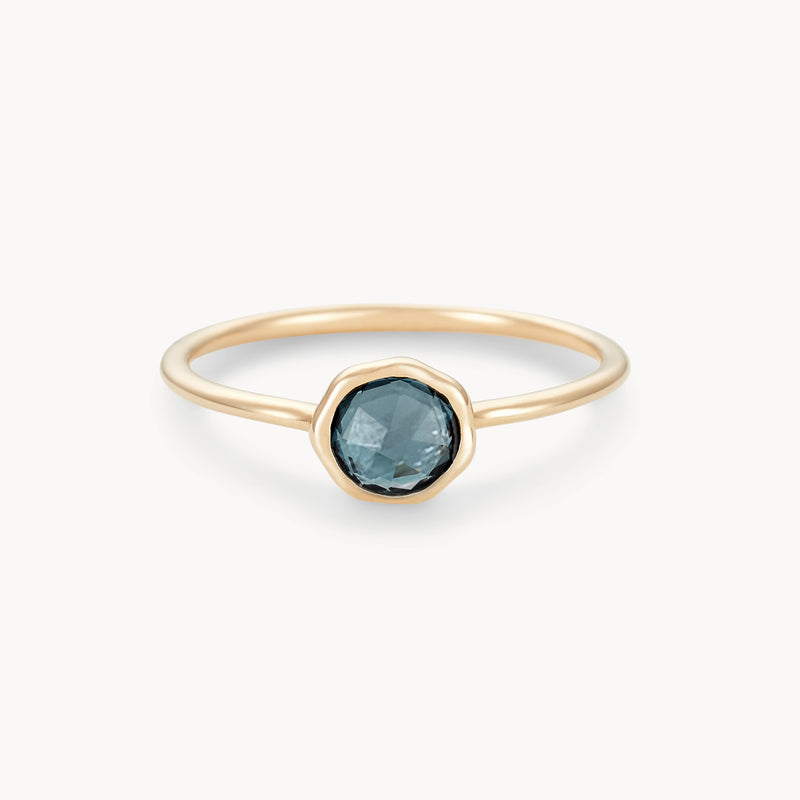 The london blue topaz mini mood ring by bluboho showcases a round, faceted dark blue topaz in a minimalist bezel setting. The thin 10k yellow gold band accentuates the simplicity and elegance of the design, providing a striking contrast to the deep hue of the gemstone. Set against a plain white background, this delicate piece radiates understated beauty.