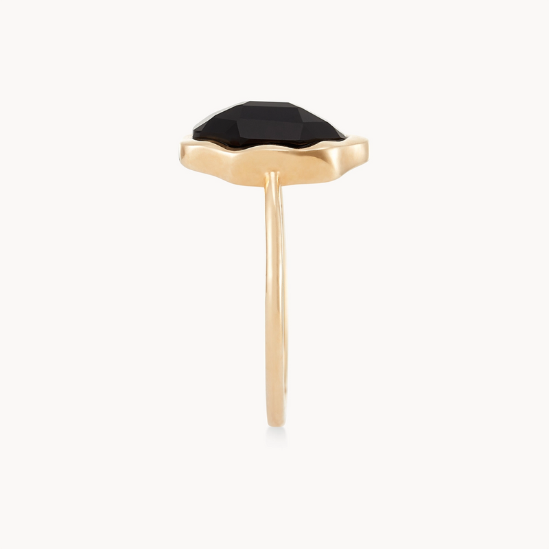 The black onyx mood ring from bluboho features a side view of a slim band made of 14k yellow gold, adorned with a black faceted gemstone in a distinctive wavy bezel setting. The gold setting secures the gemstone firmly, highlighting its dark, polished finish against the clean white backdrop.