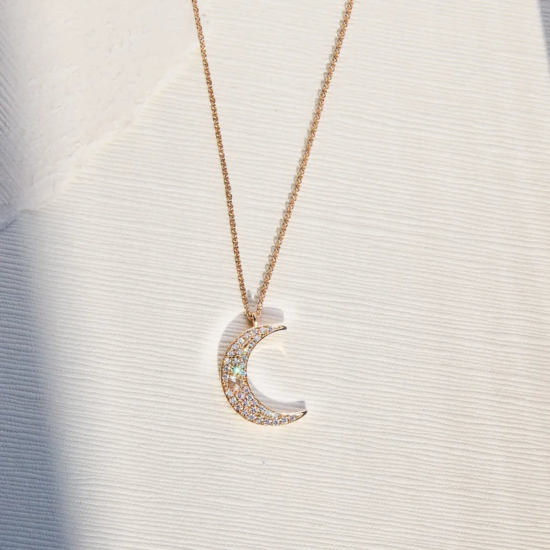 The Moonlight Crescent Diamond Necklace by bluboho features a gold chain adorned with a crescent moon pendant encrusted with small, sparkling diamonds. The pendant catches the light, enhancing its glittery appearance, and the necklace rests on a textured white fabric background.