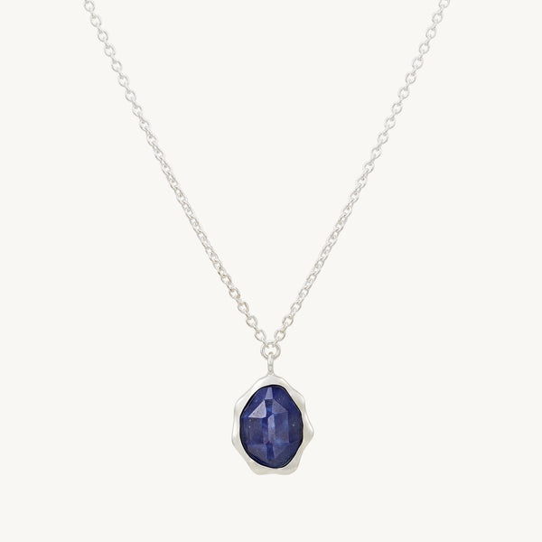 A bluboho lapis lazuli mood necklace silver - sterling silver, lapis lazuli features a thin chain and a pendant with a deep blue, faceted gemstone set in a silver bezel. The pendant has an irregular, organic shape, giving it a unique and elegant appearance. The background is white.