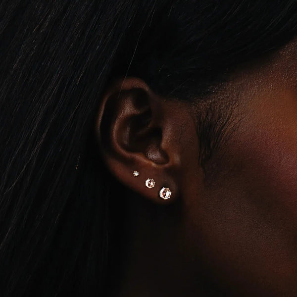 Close-up of a person's ear adorned with three pairs of earrings ascending in size. The earrings vary in design; the smallest pairs are simple studs, and the larger pairs, like the bluboho large supernova earrings, are more elaborate, featuring circular gemstones. The person's hair is straight and dark.